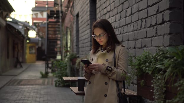 A young woman is chatting on a smartphone while standing against a wall on a narrow street. A girl in glasses and a polo with a phone in her hands. 4k