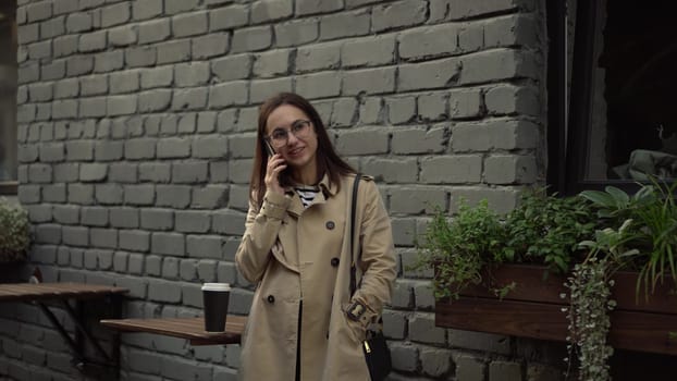 A young woman talks on the phone while standing against a wall on a narrow street. A girl with glasses and a coat speaks on the phone. 4k