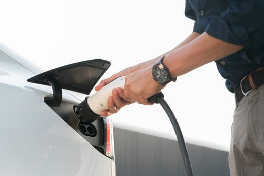 Young man put EV charger to recharge electric car's battery from charging station in city commercial parking lot. Rechargeable EV car for sustainable environmental friendly urban travel. Expedient