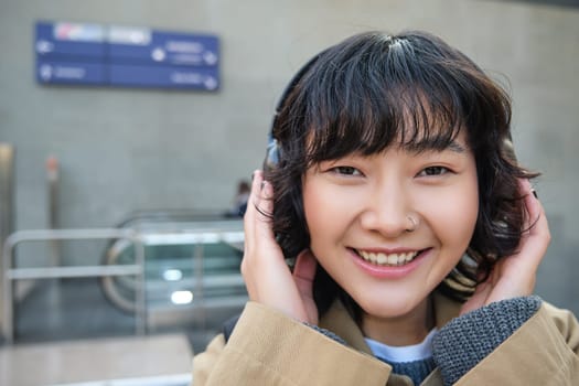 Close up portrait of stylish korean girl standing on street, listening music in headphones and smiling, posing in city centre.