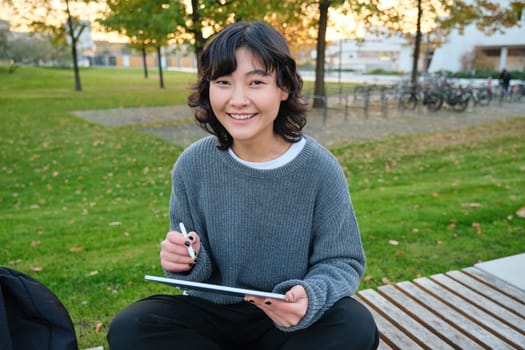 Portrait of young smiling korean girl, graphic designer, artist drawing on digital tablet with a pen tool, sitting in park on fresh air and scatching, taking notes.