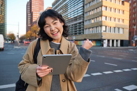 Enthusiastic asian girl with digital tablet, smiling and looking amazed, happy with something good, holding digital tablet, standing on street.