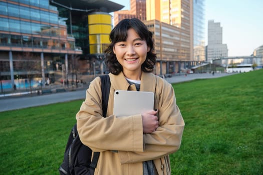 Happy asian girl stands on street, university student walks with digital tablet in hands and smiles, stands in city centre.