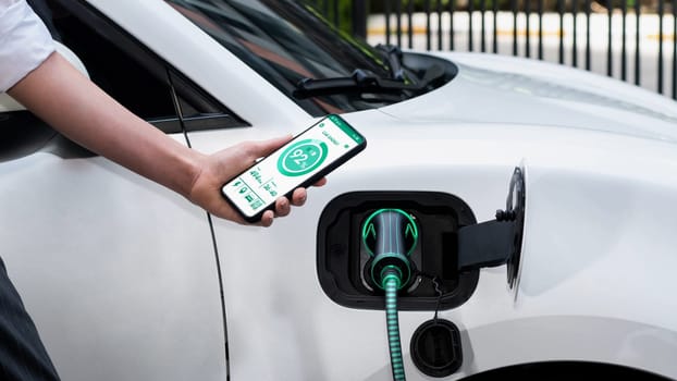 Businesswoman holding smartphone display battery status interface by smart EV mobile application while EV car recharging electricity from charging station in car park. Peruse