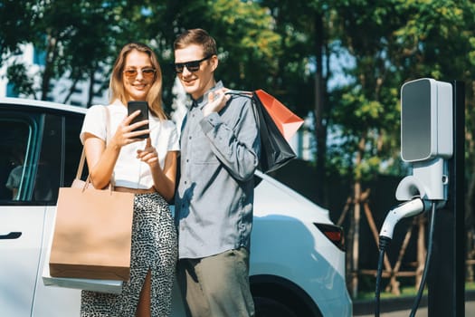 Young couple travel with EV electric car charging in green sustainable city outdoor garden in summer shows urban sustainability lifestyle by green clean rechargeable energy of electric vehicle innards