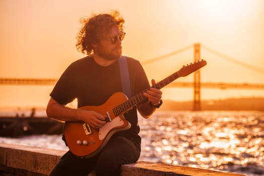 Hipster street musician in black playing electric guitar in the street on sunset on embankment with 25th of April bridge in background. Lisbon, Portugal