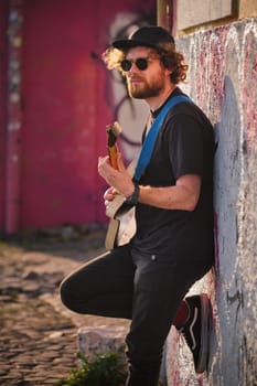 Hipster street musician in black playing electric guitar in the street on sunset leaning on a wall