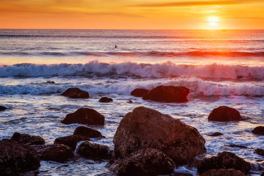 Atlantic ocean sunset with waves and rocks and surfers silhouettes in water at Costa da Caparica, Portugal