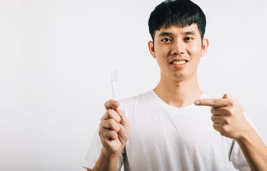 An Asian young man smiles while holding a toothbrush and brushing teeth, pointing to the brush. Studio shot isolated on white background, presenting dental health with a positive expression.