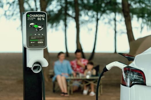 Alternative family camping trip traveling by the beach with electric car recharging battery from EV charging station with blurred family enjoying the seascape campsite background. Perpetual