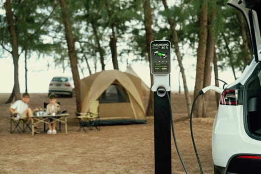Alternative family vacation trip traveling by the beach with electric car recharging battery from EV charging station with two family kid enjoying the seascape camping background. Perpetual