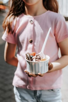Portrait of one beautiful caucasian girl holding a paper cup with ice cream, berries and cookies while standing on a city street on a clear sunny day,
