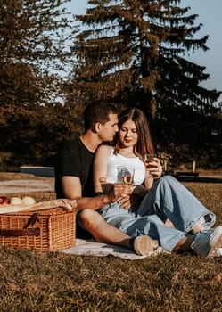 One beautiful caucasian young couple holding glasses of champagne sit hugging on a bedspread with a wicker basket and fruits in the park on a summer sunny day, close-up side view.