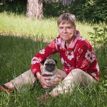 Full length view of elderly woman with dog (pug) and looking at camera sitting in grass in summer
