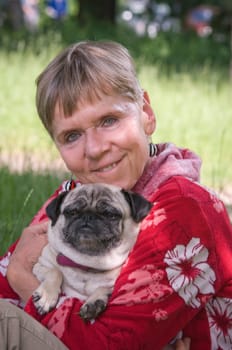 Head and shoulders portrait of mature woman in park with dog (pug) in her arms and smiling looking at camera