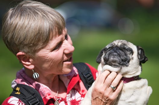 Head and shoulders view of a mature woman with dog (pug) in her arms and looking at the dog and scratching the dog's neck