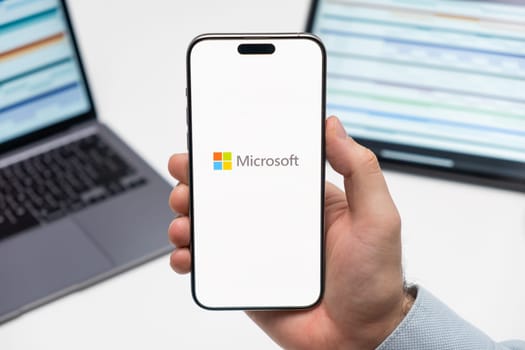 PRAGUE, CZECHIA - JULY, 2024: Microsoft logo on the screen of smartphone in mans hand and laptop on the background