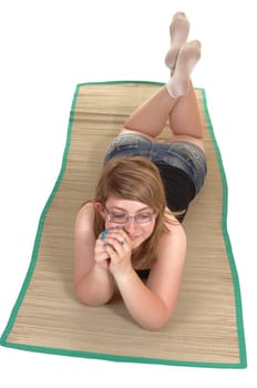 In a view from above, a teenager lying on a yoga matt on her stomach, in jeans shorts and dreaming, on white background.