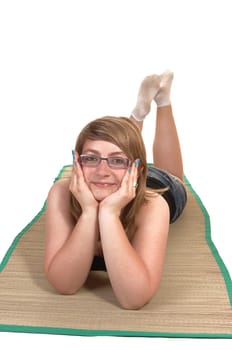 An relaxing young teenager lying on her stomach on a yoga matt on the  floor, her head in her hands and smiling into the camera, over white.