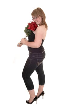 A pretty teenager standing in profile for white background, holding a bunch of red roses in her hand, in jeans and high heels.