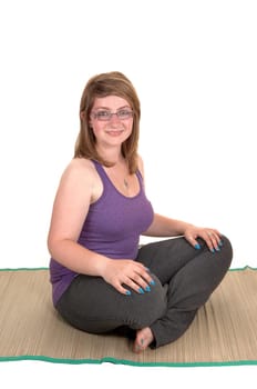 A young pretty teenager sitting on a yoga matt on the floor, with her legs grossed, smiling into the camera for white background.