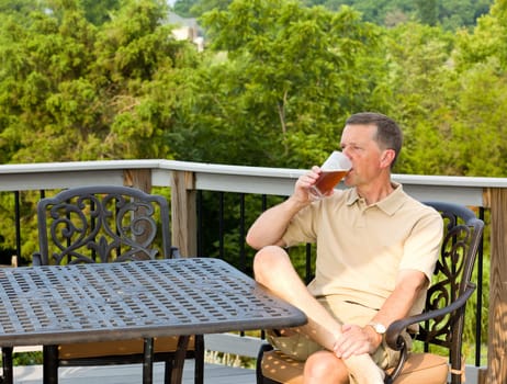 Middle aged man sitting on cast aluminium garden table on deck and drinking a glass of beer in back yard