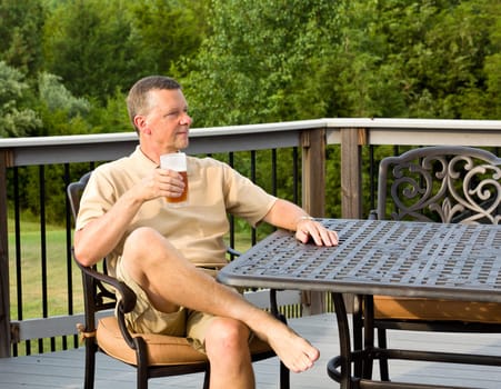 Middle aged man sitting on cast aluminium garden table on deck and drinking a glass of beer in back yard