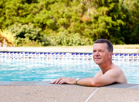 Senior male relaxing by the side of a modern swimming pool in back yard garden and facing the camera with smile