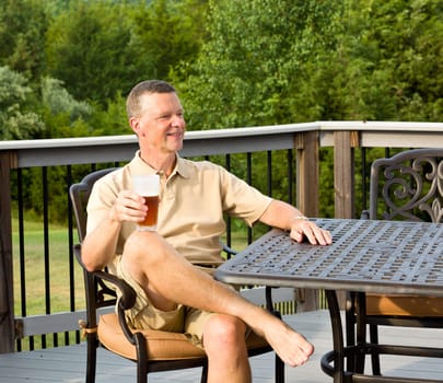 Middle aged man sitting on cast aluminium garden table on deck and drinking a glass of beer in back yard