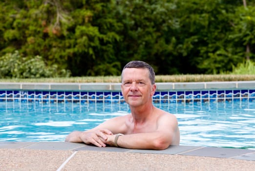 Senior male relaxing by the side of a modern swimming pool in back yard garden and facing the camera with smile