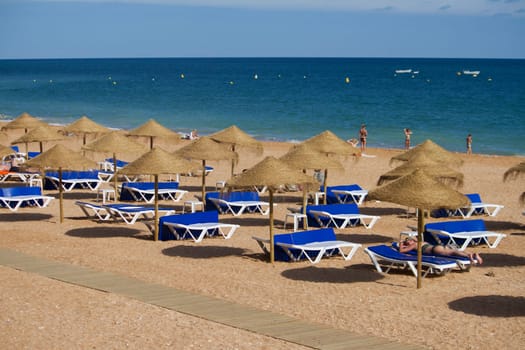 View of the beach shoreline with many straw umbrellas and blue deck chairs.