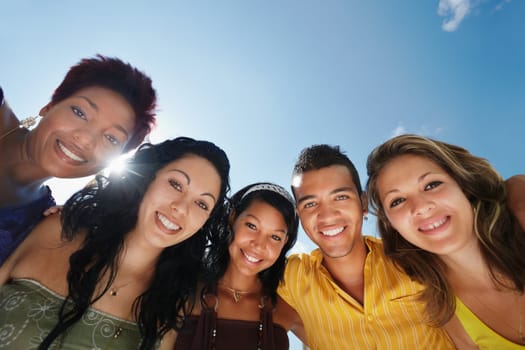 multiethnic group of five male and female friends hugging and looking at camera with sky in background. Low angle view, copy space