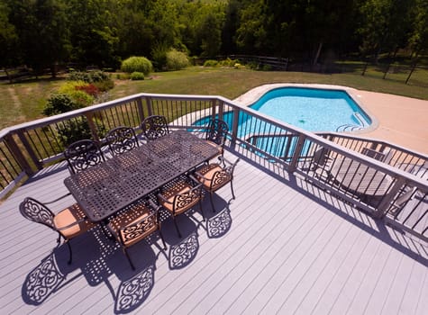 Modern table and chairs on wooden deck with swimming pool in back yard garden setting
