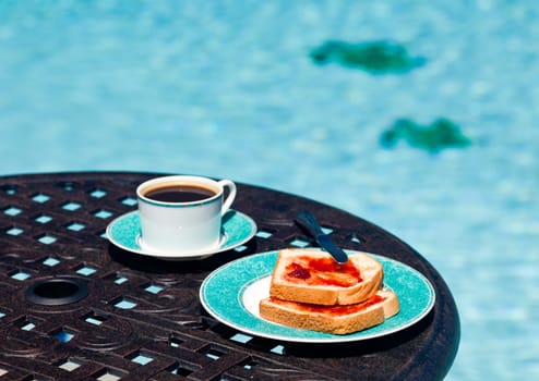 Coffee and plate on cast aluminum table and single chair by the side of swimming pool in back yard