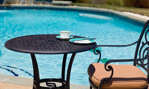 Coffee and plate on cast aluminum table and single chair by the side of swimming pool in back yard