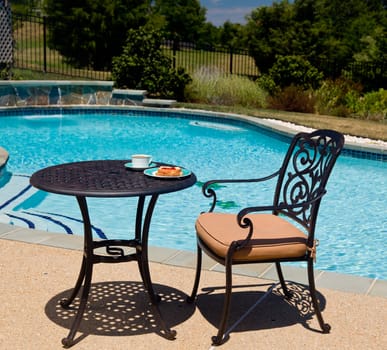 Coffee and plate on cast aluminum table and single chair by the side of swimming pool in back yard
