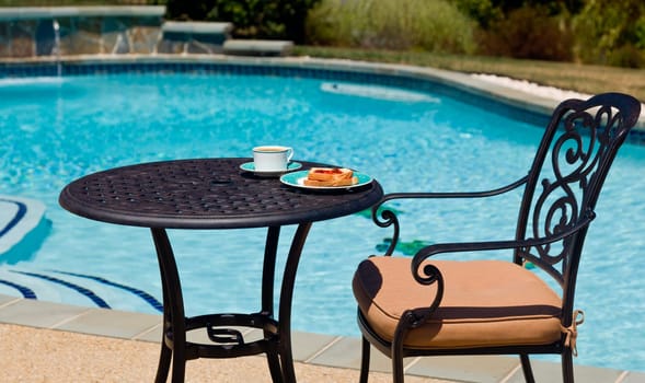 Coffee and plate on cast aluminum table and single chair by the side of swimming pool in back yard