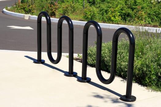 Black wavy bike rack on a sidewalk near a road in a public park on a sunny day.