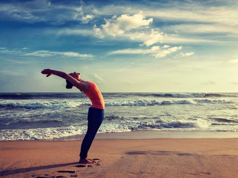 Vintage retro effect hipster style image of young sporty fit woman doing yoga Sun salutation Surya Namaskar pose Hasta Uttanasana on tropical beach on sunset