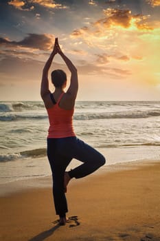 Yoga outdoors - sporty fit woman doing Hatha yoga asana Vrikshasana tre pose on tropical beach on sunset