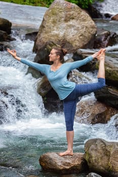 Yoga outdoors - woman doing yoga asana Natarajasana - Lord of the dance balance pose at waterfall in Himalayas