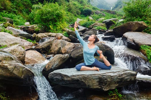 Hatha yoga outdoors - young sporty fit woman doing yoga asana Eka pada rajakapotasana - one-legged king pigeon pose at tropical waterfall. Himachal Pradesh, India