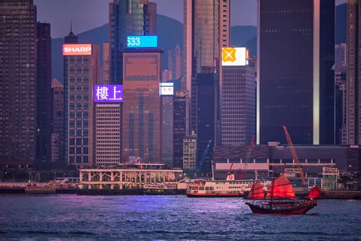 HONG KONG, CHINA - MAY 1, 2018: Tourist junk boat ferry with red sails and Hong Kong skyline cityscape downtown skyscrapers over Victoria Harbour in the evening. Hong Kong, China