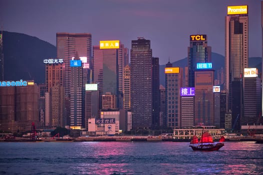 HONG KONG, CHINA - MAY 1, 2018: Tourist junk boat ferry with red sails and Hong Kong skyline cityscape downtown skyscrapers over Victoria Harbour in the evening. Hong Kong, China