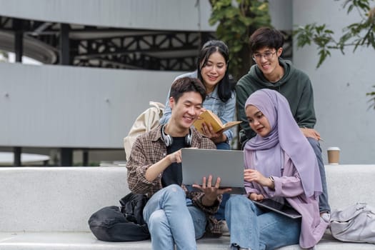 Group of happy young Asian college students sitting on a bench looking at a laptop screen, discussing and brainstorming on their school project together.