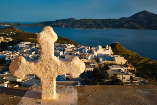 Greek Orthodox Christianity concept background - Christian cross with Plaka village on Milos island in background on sunset. Plaka town, Milos island, Greece. Focus on flowers