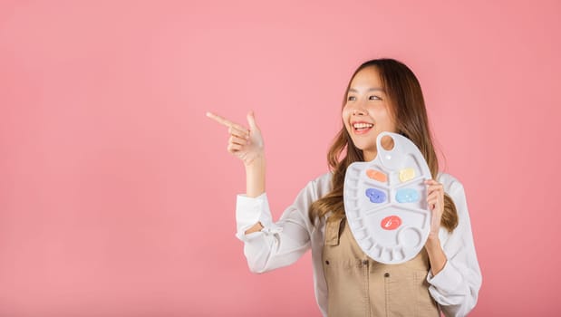 Portrait Asian young woman artist holding brush and paint palette, Happy female painting using paintbrush and palette with colors, studio shot isolated on pink background, Art design workshop