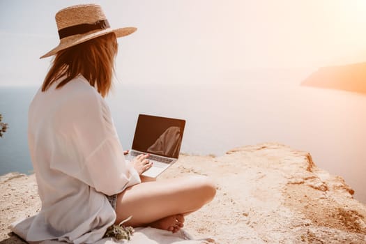 Successful business woman in yellow hat working on laptop by the sea. Pretty lady typing on computer at summer day outdoors. Freelance, travel and holidays concept.