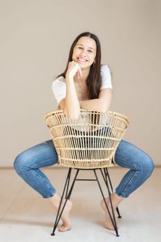 Portrait of confident beautiful woman with long brown hair, wearing casual clothes, sitting on chair in tight jeans and white t-shirt, studio background