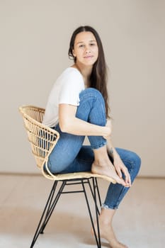 Portrait of confident beautiful woman with long brown hair, wearing casual clothes, sitting on chair in tight jeans and white t-shirt, studio background